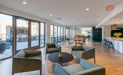 This thoughtfully designed single-floor amenity space offers a dynamic blend of social, recreational, and relaxation zones, all unified by a cohesive modern aesthetic. Light hardwood flooring runs throughout, complemented by clean white ceilings with recessed lighting that keeps the interior bright and welcoming.

The main gathering area is generously proportioned, featuring an open layout with clusters of casual seating, including a bold yellow curved sofa, vibrant orange accent chairs, and a richly patterned area rug that anchors the lounge. Large sliding glass doors and floor-to-ceiling windows flood the space with natural light and provide seamless access to an outdoor terrace furnished with bar-height seating, perfect for socializing or enjoying fresh air.

A dedicated kitchenette area offers sleek dark wood cabinetry, white quartz countertops with marble-inspired backsplash, a built-in sink, and stainless steel appliances — ideal for entertaining and community gatherings. Teal double glass doors elegantly connect this zone to the broader space.

Toward the rear, a game room with a pool table sits alongside a stylish lounge nook, where bold teal accent walls create an intimate, moody atmosphere, complemented by a pair of modern armchairs beneath a statement pendant light. An additional seating alcove features a neutral sofa and orange accent chairs against a dramatic teal backdrop, with curated artwork adding a refined finishing touch throughout.