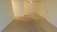 This single-floor space presents an open, unfurnished interior with a straightforward and functional design. The main living area is finished with wall-to-wall beige carpeting, providing a neutral foundation that accommodates a variety of furniture arrangements and décor styles. The walls are painted in a light, neutral tone, contributing to a bright and airy atmosphere throughout the space.

A ceiling fan is centrally mounted, offering both lighting and air circulation to keep the area comfortable year-round. The layout features an open flow, with a subtle architectural division — likely a half-wall or raised partition — separating distinct functional zones within the main living area, adding a degree of spatial definition without fully enclosing the space.

Electrical outlets are positioned along the walls at standard intervals, ensuring practical usability throughout. A door along one wall leads to an adjoining room or exterior access point, adding to the overall connectivity of the layout. The clean, move-in-ready condition of the space makes it highly adaptable, suitable for a range of living configurations. The overall ambiance is calm and neutral, offering a blank canvas for personalization.
