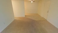 This single-floor space presents an open, unfurnished interior with a straightforward and functional design. The main living area is finished with wall-to-wall beige carpeting, providing a neutral foundation that accommodates a variety of furniture arrangements and décor styles. The walls are painted in a light, neutral tone, contributing to a bright and airy atmosphere throughout the space.

A ceiling fan is centrally mounted, offering both lighting and air circulation to keep the area comfortable year-round. The layout features an open flow, with a subtle architectural division — likely a half-wall or raised partition — separating distinct functional zones within the main living area, adding a degree of spatial definition without fully enclosing the space.

Electrical outlets are positioned along the walls at standard intervals, ensuring practical usability throughout. A door along one wall leads to an adjoining room or exterior access point, adding to the overall connectivity of the layout. The clean, move-in-ready condition of the space makes it highly adaptable, suitable for a range of living configurations. The overall ambiance is calm and neutral, offering a blank canvas for personalization.