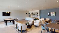 This single-floor communal amenity space is designed with a contemporary aesthetic, combining functionality and style in an open, flowing layout. Light wood-tone flooring runs throughout, unifying the various zones and contributing to a warm, inviting atmosphere.

The main lounge area features multiple seating arrangements, including plush sofas, rounded accent chairs, and low coffee tables arranged atop layered area rugs. A wall finished in a muted blue-gray tone anchors one side of the room, complemented by a wall-mounted flat-screen TV and ambient sconce lighting.

A standout dining and banquette area is defined by a textured dark wood accent wall adorned with sculptural circular wall art. A long cushioned bench seat pairs with sleek dining tables and modern side chairs, creating an ideal spot for group gatherings. Additional high-top bistro tables are positioned near floor-to-ceiling windows that offer abundant natural light and views of the surrounding greenery.

The kitchen area is a refined focal point, featuring a large white waterfall island with an undermount sink, illuminated by a row of bold black pendant lights with brass fittings. The backsplash showcases a textured tile pattern, while open shelving displays decorative accents. Warm wood cabinetry, stainless steel appliances, and a full-size refrigerator complete the well-appointed kitchen. Recessed lighting throughout ensures the entire space remains bright and welcoming.
