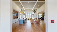 Located at 100 Plyer Cir in Indian Trail, NC, this thoughtfully designed community clubhouse offers a generous and well-appointed single-floor layout. The space immediately impresses with its soaring coffered ceilings accented by gray shiplap paneling, warm wood-look tile flooring, and an abundance of natural light streaming through floor-to-ceiling windows that frame views of the outdoor pool area.

The open main gathering area serves as the social heart of the space, featuring a central kitchen island with a bold blue base and white countertop, bar seating, and a textured accent wall with an integrated television — ideal for community events. Statement pendant lighting and a dramatic sculptural chandelier add visual depth overhead.

Adjacent lounge areas offer carefully curated seating arrangements with plush accent chairs, layered rugs, and curated artwork throughout, creating an inviting atmosphere for relaxation or conversation. A shiplap-walled nook provides a more intimate setting with a wall-mounted TV.

A bright corridor connects to a dedicated workspace and business services area, complete with small café-style tables, workstation counters, and office equipment. Colorful roman shades and warm pendant lighting give this corridor a welcoming, boutique feel. Throughout, the design balances modern finishes with warm, eclectic touches for a refined yet approachable ambiance.