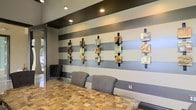 Located at 471 N Hamilton Rd in Gahanna, Ohio, this single-story space presents a polished, contemporary interior that blends artistic detail with functional design. The space immediately draws attention to its bold accent wall, finished in wide horizontal stripes of contrasting gray tones that create a strong visual focal point. Mounted along this wall is a curated collection of mixed-media art pieces, each displayed on vertical black metal supports, adding depth, color, and a gallery-like quality to the environment.

The flooring features a distinctive mosaic-style tile in warm, earthy tones, providing a rich textural contrast to the cooler gray palette of the walls. Seating includes modern gray upholstered chairs positioned at a sleek glass-top table, contributing to a clean and professional atmosphere. Recessed lighting and track lighting fixtures installed in the ceiling provide layered illumination, highlighting both the artwork and the overall layout of the space.

The overall ambiance is modern and refined, with thoughtful design choices that balance aesthetics and comfort. The combination of artistic wall displays, quality materials, and contemporary furnishings gives the space a sophisticated character well-suited for professional or creative use.