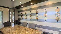 Located at 471 N Hamilton Rd in Gahanna, Ohio, this single-story space presents a polished, contemporary interior that blends artistic detail with functional design. The space immediately draws attention to its bold accent wall, finished in wide horizontal stripes of contrasting gray tones that create a strong visual focal point. Mounted along this wall is a curated collection of mixed-media art pieces, each displayed on vertical black metal supports, adding depth, color, and a gallery-like quality to the environment.

The flooring features a distinctive mosaic-style tile in warm, earthy tones, providing a rich textural contrast to the cooler gray palette of the walls. Seating includes modern gray upholstered chairs positioned at a sleek glass-top table, contributing to a clean and professional atmosphere. Recessed lighting and track lighting fixtures installed in the ceiling provide layered illumination, highlighting both the artwork and the overall layout of the space.

The overall ambiance is modern and refined, with thoughtful design choices that balance aesthetics and comfort. The combination of artistic wall displays, quality materials, and contemporary furnishings gives the space a sophisticated character well-suited for professional or creative use.
