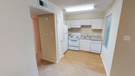 This single-floor unit offers a practical and efficient layout designed to maximize its compact footprint. The kitchen is outfitted with white cabinetry extending to the upper walls, providing ample storage space. A full suite of white appliances includes a refrigerator, electric coil range with oven, dishwasher, and a single-basin stainless steel sink set into a speckled laminate countertop. Light wood-look flooring runs throughout the kitchen, creating a clean, easy-to-maintain surface. A ventilation hood is mounted above the range, and a ceiling-mounted fluorescent light fixture provides functional illumination.

The living area and bedroom both feature neutral-toned carpet and ceiling fans with integrated light fixtures, contributing to a consistent, understated aesthetic throughout. Textured popcorn ceilings are present in all rooms. Warm beige walls lend a cohesive tone across the unit.

The bathroom includes a full tub-shower combination with tiled surround, a toilet, and a single vanity with a speckled laminate countertop, white basin sink, and a large mirror topped by a row of globe lights. The overall design is straightforward and functional, suited for efficient everyday living.