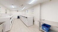 Located at 59 Concession St in Cambridge, Ontario, this shared laundry facility is a well-organized, single-floor space designed for efficient, high-volume use. The room is laid out to maximize functionality, with rows of front-loading washing machines and dryers lining both sides, providing ample capacity for building residents. The flooring features a crisp herringbone-patterned tile in a light neutral tone, lending a clean and modern aesthetic to the utilitarian space. Bright fluorescent strip lighting runs along the ceiling, ensuring the room is well-lit at all times. The walls are finished in a clean white, contributing to an open and sanitary atmosphere. A stainless steel utility sink is conveniently positioned near the entrance, alongside a stainless steel folding table that provides a practical surface for sorting and organizing laundry. A payment kiosk station is visible, indicating a card or coin-operated system for machine use. A rolling laundry hamper cart and a recycling bin are thoughtfully provided for user convenience. Instructional signage is posted on the walls, offering guidance on proper washer and dryer operation. The space is accessed through a secure door and includes a floor drain, reflecting a practical, well-maintained design built for everyday communal use.