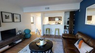 Located at 300 E Evans Street in the heart of West Chester, PA, this single-floor residence offers a thoughtfully designed open-concept layout that seamlessly connects the living and kitchen areas. The living room is welcoming and well-lit, featuring warm hardwood flooring, neutral white walls, and ample space for comfortable furnishings. A round accent table and area rug anchor the seating area, creating an inviting focal point within the room. Built-in shelving adds functional storage and display space along one wall, complementing the room's clean aesthetic. The kitchen is positioned at the heart of the home, accessible via a pass-through bar counter with room for bar stool seating — ideal for casual dining or entertaining. Dark espresso cabinetry, stainless steel appliances including a microwave and range, and light countertops give the kitchen a modern, polished look. A hallway visible from the living area leads to additional spaces beyond, with a closet offering convenient storage. The consistent hardwood flooring flows throughout, unifying the space with warmth and continuity. Overall, the residence balances a modern, functional design with a cozy and approachable ambiance, making it a practical and attractive home in a prime West Chester location.