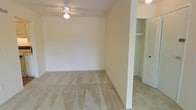 This single-floor space presents a straightforward and functional layout with clean, neutral interiors throughout. The main room is finished with wall-to-wall beige carpeting that lends a soft, comfortable feel underfoot, while the white walls provide a bright, blank canvas well-suited to a variety of furnishing styles. A ceiling fan with an integrated light fixture is centrally mounted, offering both overhead lighting and air circulation. The room is adequately sized to accommodate standard bedroom or living area furniture arrangements. Adjacent to the main room, a structural column marks the transition to a secondary area, also carpeted, which leads to a closet outfitted with louvered bifold doors and a built-in shelf above — providing practical everyday storage. A doorway visible in the background hints at additional rooms or connecting spaces beyond. The overall ambiance is clean, understated, and move-in ready, with a neutral palette throughout that makes the space highly adaptable. The absence of clutter and the freshly maintained condition of the carpeting and walls suggest a well-kept interior prepared for new occupancy.