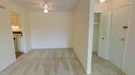 This single-floor space presents a straightforward and functional layout with clean, neutral interiors throughout. The main room is finished with wall-to-wall beige carpeting that lends a soft, comfortable feel underfoot, while the white walls provide a bright, blank canvas well-suited to a variety of furnishing styles. A ceiling fan with an integrated light fixture is centrally mounted, offering both overhead lighting and air circulation. The room is adequately sized to accommodate standard bedroom or living area furniture arrangements. Adjacent to the main room, a structural column marks the transition to a secondary area, also carpeted, which leads to a closet outfitted with louvered bifold doors and a built-in shelf above — providing practical everyday storage. A doorway visible in the background hints at additional rooms or connecting spaces beyond. The overall ambiance is clean, understated, and move-in ready, with a neutral palette throughout that makes the space highly adaptable. The absence of clutter and the freshly maintained condition of the carpeting and walls suggest a well-kept interior prepared for new occupancy.