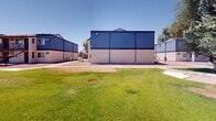 Located at 1829 North 45th Avenue in Phoenix, Arizona, this single-level outdoor community space is designed to serve as a central recreational hub for its surrounding residential neighborhood. The centerpiece of the grounds is a full-sized basketball court with clearly marked lines and regulation hoops, providing ample space for both casual play and organized games. Surrounding the court, expansive stretches of well-maintained green lawn offer open space for leisure, gatherings, and informal activities. Towering palm trees are scattered throughout the grounds, adding a distinctly Southwestern character while offering patches of natural shade under the bright Arizona sun. A dedicated children's playground, complete with play structures and a shaded canopy, is visible within the grounds, making the space family-friendly and accessible to residents of all ages. The broader campus includes several single-story and two-story buildings with blue metal-paneled facades, contributing a utilitarian yet cohesive architectural style. Additional landscaping features mature olive and shade trees that provide natural canopy coverage in certain areas. The entire outdoor environment benefits from the clear desert skies and open sightlines, creating a bright, active, and community-oriented atmosphere well suited for year-round outdoor recreation in the Phoenix climate.