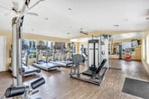 Located at 24850 Hancock Ave in Murrieta, California, this fitness center offers a generously sized, single-floor exercise room designed to accommodate a full range of workout routines. The space features warm yellow walls, wood-style flooring, and white ceilings fitted with recessed lighting and a ceiling fan, creating a bright and energizing atmosphere.

The room is comprehensively equipped for both cardio and strength training. Cardio equipment includes multiple treadmills and stationary bikes, each outfitted with individual monitors. For strength training, the space provides Life Fitness cable and functional training machines, a leg press, adjustable weight benches, and a well-stocked dumbbell rack displayed alongside a large wall mirror that enhances the sense of openness. Medicine balls and exercise balls are neatly arranged for functional and core training.

Natural light streams in through several windows positioned along the walls, some of which offer views of an outdoor area. Glass-paned doors provide additional access and light. A wall-mounted flat-screen television adds an entertainment element to support longer workout sessions. The open floor plan allows for comfortable movement between stations, making this fitness center a practical and inviting space for a comprehensive exercise experience.