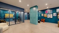Located at 2061 Rue Stanley in downtown Montréal, this striking lobby serves as the welcoming centerpiece of the Loxley building. The space makes an immediate impression with its bold, curated color palette — deep navy blue paneled walls contrast beautifully with teal-painted structural columns and millwork, creating a cohesive and vibrant aesthetic throughout.

The flooring consists of large-format light beige tiles that span the generous open area, lending a clean and polished foundation to the dynamic design above. A glass-enclosed lounge area sits to one side, offering a semi-private seating zone furnished with modern chairs and a work table, ideal for informal meetings or focused work.

Throughout the lobby, multiple seating vignettes are thoughtfully arranged, featuring contemporary furniture including rounded cream sofas, an orange lounge chair, and earthy-toned accent pieces. Curated artwork lines the teal-accented feature wall, adding color and cultural character to the space.

One corridor wall showcases a dramatic dark grey exposed brick surface, adorned with a large, colorful sculptural installation that commands attention and adds an artistic focal point. Additional wall-mounted sculptural pieces further reinforce the lobby's gallery-like quality.

Floor-to-ceiling windows along the entrance flood the space with natural light and offer views of the surrounding streetscape, enhancing the open and inviting atmosphere of this thoughtfully designed lobby.