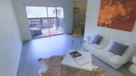This single-floor space showcases a clean, modern aesthetic defined by an open-plan layout and an abundance of natural light. The living area serves as the heart of the space, featuring light hardwood-style flooring, crisp white walls, and a contemporary arrangement that includes a white sofa accented with grey throw pillows, a sleek white coffee table, and a distinctive cowhide rug that adds organic texture to the room. A bold abstract artwork in warm orange tones provides a striking focal point on one wall.

Large sliding glass doors form a seamless transition between the interior living area and an outdoor deck, which is fitted with horizontal railings and features a red accent mat, allowing for excellent natural light penetration and effortless indoor-outdoor living. Views of surrounding greenery are visible beyond the deck.

Adjacent to the living area, a dining nook accommodates a compact round table with minimalist chairs in a muted yellow tone, maintaining the contemporary and uncluttered character of the space. A glimpse into a secondary room reveals additional functional space with simple, neutral finishes. Throughout, the design prioritizes openness, light, and modern simplicity.