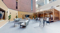 The 77 West outdoor terrace is a thoughtfully designed urban amenity space nestled within a courtyard framed by the building's distinctive brick exterior and warm wood-panel accent walls. The terrace is organized into several distinct zones, each catering to a different style of outdoor enjoyment. A central lounge area features a generously sized sectional sofa and armchairs with neutral cushions set around low coffee tables, creating a comfortable gathering spot for relaxed conversation. An outdoor kitchen station is equipped with a full-sized gas grill and ample counter space, making it ideal for resident cookouts and social events. A wall-mounted television with barn-door shutters serves as a focal point for outdoor entertainment. High-top tables and bar-height chairs are distributed throughout the terrace, offering casual dining and social seating options. A raised pergola structure with a timber frame and canopy provides a shaded retreat with additional high-top seating, accessible via wood-and-steel steps with a modern railing. Wood benches and additional bistro sets along the perimeter complete the space. The open-air design offers views of the surrounding downtown skyline, giving the terrace a vibrant urban atmosphere while maintaining a comfortable and inviting environment.