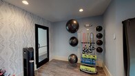 Located at 106 Pullman Lane in Mooresville, NC, this single-level fitness center is thoughtfully designed to support a comprehensive workout experience. The main exercise floor is spacious and open, finished with durable rubber flooring and featuring large black-framed windows that invite abundant natural light and views of the surrounding community. Ceiling fans overhead contribute to a comfortable workout environment, while a wall-mounted television provides entertainment during training sessions.

The strength training area is well-appointed with a full rack of dumbbells, a power rack, weight benches, and cable machines. Large wood-paneled accent walls add warmth and contrast to the otherwise crisp, neutral interior. Floor-to-ceiling mirrors are strategically placed to allow users to monitor their form throughout their workout.

The cardio section houses a lineup of treadmills, upright and recumbent bikes, and elliptical machines, all positioned to face the exterior windows. A dedicated functional training room, accessible through glass-paneled doors, features textured accent wallpaper, wall-mounted stability balls, resistance bands, medicine balls, and foam rollers neatly organized on a multi-tier rack. The overall ambiance is modern and professional, balancing aesthetics with practical fitness functionality.