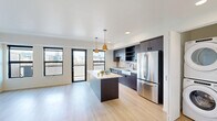 Located in the vibrant SE Belmont corridor of Portland, Oregon, this thoughtfully designed one-bedroom, one-bathroom unit offers a contemporary living experience with high-end finishes and an efficient, open-concept floor plan. Light hardwood flooring flows throughout the space, complementing the clean white walls and modern aesthetic.

The open living and kitchen area serves as the heart of the unit, bathed in natural light from a generous bank of black-framed windows that frame striking views of the Portland skyline. A sliding glass door provides access to a private balcony. The kitchen is a standout feature, equipped with dark espresso cabinetry, a hexagonal tile backsplash, quartz countertops, stainless steel appliances including a French door refrigerator and built-in oven, a farmhouse-style sink, and a central island with pendant lighting overhead. A stacked washer and dryer unit is conveniently integrated into the space.

The bedroom is bright and well-proportioned, with dual city-view windows, a wall-mounted climate control unit, and direct access to a spacious walk-in closet with built-in shelving. The bathroom features dark cabinetry with an undermount sink, matte black fixtures, and a full tub-shower combination with clean white tile surround.