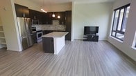 This contemporary single-floor space showcases a thoughtfully designed open-concept layout that seamlessly integrates the kitchen and living area into one cohesive environment. The kitchen is a standout feature, outfitted with rich, dark espresso cabinetry that extends to the upper walls, providing ample storage while lending a bold, modern aesthetic. A mosaic tile backsplash in warm earth tones adds texture and visual interest between the upper and lower cabinets. The kitchen is equipped with stainless steel appliances, including a range, microwave, and refrigerator, complemented by a central island topped with a light granite surface — ideal for meal preparation or casual dining. Pendant lighting above the island and a track light fixture add layered illumination to the kitchen zone. The open living area flows naturally from the kitchen, unified by wide-plank, light grey wood-look flooring that runs throughout the space, creating a clean and airy feel. Neutral-toned walls enhance the brightness of the interior, while the generous floor plan allows for flexible furniture arrangements. The overall ambiance is modern and functional, balancing dark design accents with light, open surroundings for a well-grounded yet contemporary atmosphere.
