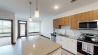 This thoughtfully designed single-floor residence offers a clean, contemporary aesthetic throughout. The heart of the home is an open-concept kitchen and living area, anchored by a generously sized center island with a quartz-style countertop — ideal for dining or meal prep. The kitchen is outfitted with stainless steel appliances including a refrigerator, range, microwave, and dishwasher. Upper cabinetry features a warm wood finish, while lower cabinets are rendered in crisp white, creating a two-tone contrast that adds visual depth. A classic white subway tile backsplash and recessed lighting, complemented by stylish pendant lights above the island, complete the kitchen's polished look. Light wood-tone flooring runs throughout the main living areas, enhancing the bright and airy atmosphere. Natural light flows in through large windows fitted with horizontal blinds. The bathroom is well-appointed with a full soaking tub and shower combination, a vanity with storage, and durable flooring. The bedroom is a calm, comfortable retreat featuring plush carpeting, a ceiling fan, and a wide window offering outdoor views. A dedicated laundry area with wall-mounted storage adds functional convenience to this well-rounded space.