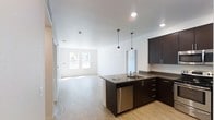 This single-floor space showcases a contemporary open-concept layout that seamlessly connects the kitchen and living area. The kitchen is outfitted with rich dark cabinetry that provides a striking contrast against the white walls and light hardwood flooring that extends throughout the unit. A peninsula with a built-in sink and stainless steel dishwasher serves as a natural divider between the kitchen and living space, offering both functionality and visual definition. Sleek pendant lights hang above the peninsula, adding focused task lighting while contributing to the modern aesthetic. Dark countertops complement the cabinetry and carry through the kitchen's cohesive design. Recessed lighting in the ceiling provides even, ambient illumination across the open space. The living area benefits from natural light streaming through a window along the far wall, creating a bright and airy atmosphere. The clean lines, neutral color palette, and high-quality finishes throughout give the space a polished, move-in-ready feel, well-suited for a range of lifestyles.