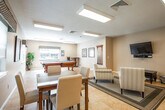 Located at 500 Peconic St in Ronkonkoma, NY, this single-floor community amenity space is designed to offer a welcoming and versatile environment for recreation and relaxation. The layout flows openly across several functional zones, all unified by warm beige tones, neutral walls, and light-toned tile flooring throughout.

The standout feature is a dedicated billiards area, where a full-size pool table with a vibrant blue felt surface takes center stage. This area is brightened by natural light from windows dressed with stylish patterned valances, and a wall-mounted cue rack keeps equipment neatly accessible. A cozy high-top table with bar seating and framed artwork on the walls add character to the space.

Adjacent to the billiards zone is a comfortable lounge area furnished with striped upholstered armchairs arranged around a dark wood coffee table, all set atop an area rug. A large wall-mounted flat-screen TV anchors this seating area, creating an ideal spot for gathering and entertainment.

A separate dining area provides a more intimate setting, featuring a glass-top dining table with upholstered chairs and a window offering views of the surrounding greenery. Recessed and overhead fluorescent lighting ensures the entire space is well-lit throughout the day. A service window and entry door connect the space to the broader facility, enhancing its functionality as a shared community hub.