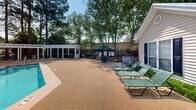 Located at 930 E Main St in Lexington, South Carolina, this outdoor amenity area is a well-appointed community recreational space designed for relaxation and leisure. At its centerpiece is a generously sized swimming pool with clearly marked depth indicators, surrounded by a textured, slip-resistant aggregate deck that provides ample space for sunbathing and socializing. Multiple green lounge chairs are positioned poolside, offering comfortable spots to unwind under the South Carolina sun.

A striking white pergola structure runs along one side of the pool area, providing shaded seating beneath its open-beam canopy — an ideal retreat from the heat. Additional outdoor dining and seating areas, shaded by green umbrellas, are situated nearby for casual gatherings. A children's playground is visible in the background, adding a family-friendly dimension to the space.

The pool area is enclosed by a secure fence and is framed by a lush canopy of mature trees, including tall pines, that provide natural shade and privacy. Adjacent to the pool deck stands a charming single-story building featuring a distinctive arched, multi-pane window facade and a metal roof, which likely serves as a clubhouse or community facility. The overall ambiance is inviting, serene, and well-maintained, making it an excellent outdoor retreat.