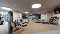 This fitness center is a thoughtfully designed, single-floor facility offering a comprehensive workout environment for users of all fitness levels. The space is organized into distinct functional zones, creating an efficient and intuitive layout throughout.

The cardio zone features a row of treadmills and an elliptical trainer positioned along floor-to-ceiling windows, flooding the area with natural light and offering views of the surrounding outdoor environment. Large, circular pendant lights complement the recessed lighting overhead, creating a bright and energizing atmosphere.

The strength training area is well-appointed with a selection of Precor cable and resistance machines, a dedicated free weights section with a tiered dumbbell rack, and flat workout benches. Full-length mirrors line the walls, enhancing the sense of openness while allowing users to monitor their form.

A dedicated functional training zone features rubber flooring tiles in a two-tone pattern, a heavy tire for tire flips, stability balls, medicine balls, and TRX suspension trainers anchored to a wall-mounted rig. A flat-screen television is mounted on the wall for added convenience.

The entrance area provides a welcoming transition into the facility, complete with cubby storage, colorful wall art, and posted fitness center guidelines. The durable, cushioned flooring throughout the space ensures comfort and safety during all types of workouts.