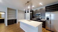 This single-floor space showcases a contemporary design with a thoughtfully appointed open-concept layout. At the heart of the home is a striking kitchen, anchored by a spacious white quartz island with an undermount sink, offering generous prep and seating space. Dark espresso cabinetry extends to the ceiling, providing ample storage, while a mosaic tile backsplash adds texture and visual interest. Stainless steel appliances, including a range and over-the-range microwave, complete the modern aesthetic. Three sleek black pendant lights hang above the island, creating a bold focal point and warm task lighting. Recessed ceiling lights throughout ensure the space remains bright and well-illuminated. Light wood-tone flooring runs continuously through the open area, adding warmth and cohesion. Adjacent to the kitchen, built-in open shelving and a set of paneled doors offer versatile storage options, while a compact beverage cooler built into the cabinetry adds a refined convenience. The overall ambiance is clean, modern, and functional, with a neutral color palette of white, grey, and dark wood tones that lend the space a sophisticated yet welcoming character.