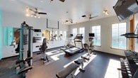 This fitness center is a fully equipped, single-floor gym facility designed to support a comprehensive workout routine. The space is divided into distinct functional zones, seamlessly connected under consistent ceiling heights with multiple ceiling fans providing continuous airflow throughout. The cardio area is bathed in natural light from a row of large windows, accommodating treadmills, an elliptical, and a stationary bike, each outfitted with individual monitor screens. A full-length mirrored wall runs along one side, visually expanding the space while allowing users to monitor their form. The strength training section features a multi-station cable machine, an adjustable workout bench, and a well-stocked dumbbell rack. A wall-mounted flat-screen television is positioned for convenient viewing during workouts. The flooring throughout consists of durable rubber gym matting, ideal for high-impact activity. Bold teal accent walls introduce a vibrant, energizing atmosphere, contrasting with the neutral white and gray tones of the surrounding walls. Track lighting supplements natural light in the interior zones. A water cooler station and additional wooden cabinetry offer practical amenities, completing a thoughtfully designed fitness space suited for a variety of training needs.