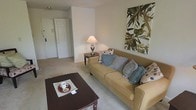 This single-floor space presents a welcoming living area designed with comfort and functionality in mind. The lounge is finished with wall-to-wall carpeting in a soft neutral tone, contributing to a warm and relaxed atmosphere throughout. A generously sized tan sofa serves as the focal point of the room, dressed with an assortment of decorative blue and patterned throw pillows that add texture and subtle color contrast. A dark wood coffee table anchors the seating area, styled simply with a tray and cup, reinforcing a tidy, livable aesthetic. Adjacent to the sofa, a dark wood end table supports a classic table lamp, providing warm ambient lighting. A large botanical wall print adds visual interest to the otherwise understated wall space, introducing a natural element to the interior. Neutral beige walls keep the space feeling open and bright. A doorway visible in the background leads further into the unit, hinting at additional rooms beyond the living area. Closet doors near the entry suggest practical storage solutions. Overall, the space offers a clean, unpretentious layout suited to everyday living.