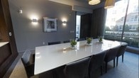 This single-floor space presents a polished and contemporary interior designed for meetings or formal dining. At its centerpiece sits a large, sleek white oval conference or dining table surrounded by cushioned dark charcoal chairs, comfortably accommodating a sizable group. The table is elegantly dressed with glass bowl centerpieces containing green botanical arrangements, adding a touch of nature to the refined setting.

The walls are finished in a sophisticated medium grey tone, complemented by warm wood-framed wall sconces that cast a soft, directed light across the room. A framed piece of abstract artwork adorns one wall, contributing to the curated aesthetic. Overhead, striking yellow-gold pendant lights provide both functional illumination and a bold design accent against the neutral palette.

Natural light filters into the space through large windows, offering views of outdoor greenery and the surrounding streetscape. A structural column with an industrial character adds architectural interest near the center of the room. Dark wood paneling and curtains frame a secondary opening, enhancing the sense of depth and warmth. The overall ambiance is calm, professional, and welcoming — a well-appointed space suited for productive gatherings or upscale entertaining.