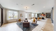 Located at 2880 Avila Cross Cir in Oviedo, Florida, this contemporary community living space is thoughtfully designed to accommodate a variety of social activities across a single, expansive floor. The interior is unified by light-toned wood-look tile flooring and crisp white walls, creating a bright and welcoming atmosphere throughout.

The main lounge area is anchored by a plush sectional sofa, a sculptural coffee table, and bold pendant lighting with an organic, branching design. Large windows dressed with linen drapes flood the space with natural light and offer views of the surrounding community. Adjacent to the lounge, a casual seating cluster features modern accent chairs in neutral tones accented with mustard yellow pillows, arranged around a patterned area rug.

The entertainment zone transitions seamlessly into a dedicated gaming area, complete with a poker table surrounded by rolling leather chairs, a pool table, and a shuffleboard table set on plush carpeting. A wall-mounted fireplace display and built-in shelving serve as a stylish focal point in this section. A work or dining area outfitted with a dark table and leather task chairs provides a more focused setting.

Geometric wall panels and contrasting gray accent walls add architectural interest throughout, while recessed lighting and designer pendants ensure a well-lit, polished ambiance. A sleekly finished stairwell with textured wallcovering connects to additional levels of the building.