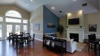 Located at 2590 Woodland Dr in New Bern, NC, this single-floor space makes a strong impression with its open, flowing layout and thoughtfully designed interior. Soaring vaulted ceilings amplify the sense of space, drawing the eye upward and creating an airy, expansive atmosphere throughout. Rich hardwood flooring runs underfoot, adding warmth and a timeless elegance to the entire area.

A striking teal-blue accent wall serves as a bold architectural focal point, rising dramatically to meet the vaulted ceiling and visually anchoring the living area. The living space features a classic white-framed fireplace flanked by a wall-mounted flat-screen television, creating an inviting and functional gathering area. Comfortable seating arrangements and a decorative console table complete the living zone.

Adjacent to the living area, a defined dining space accommodates a dark wood table and chairs, offering an ideal setting for meals and conversation. Large windows along one wall flood the space with abundant natural light, offering pleasant outdoor views and enhancing the open, cheerful ambiance. Recessed lighting throughout provides additional illumination. The cohesive neutral wall tones, accented by the bold feature wall, tie the space together into a harmonious and welcoming environment.