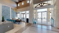 This residential community showcases a thoughtfully designed clubhouse and outdoor amenity area that blends modern and rustic architectural elements. The clubhouse interior is defined by soaring ceilings with exposed timber beams, prominent limestone columns, and floor-to-ceiling glass doors that flood the space with natural light. Light wood flooring in a herringbone pattern anchors the interior, while a dining area with wooden tables and teal accent chairs adds warmth and color. Decorative blue glass vases and an artistic wall feature contribute to the polished aesthetic. Large glass doors open directly to the outdoor amenity area, creating a seamless indoor-outdoor connection.

Outside, a generously sized resort-style pool takes center stage, featuring crystal-clear blue water, sunning shelves, and ample lounge seating. The pool deck is bordered by a covered pavilion with dramatic exposed wood trusses and iron hardware set atop stone pillars, providing shade over a built-in bar counter and lounge seating area. The surrounding multi-story residential buildings, finished in neutral tones with stone accents and private balconies, frame the outdoor space, creating an enclosed and cohesive community atmosphere.