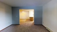 This single-story space presents a straightforward, functional layout with a clear flow between living areas. The main room features wall-to-wall neutral carpet and is painted in a calming blue-gray tone, creating a subdued, cohesive atmosphere. A wide doorway opening connects this area to an adjoining room, enhancing the sense of openness and allowing natural sightlines throughout the space.

The adjoining room, visible through the passthrough opening, is finished in a lighter, warm white tone and is equipped with a ceiling fan, providing both ventilation and ambient lighting. A wooden cabinet or kitchen island is partially visible near the transition point between the two areas, suggesting proximity to a kitchen or dining space.

Throughout both areas, white baseboards provide clean contrast against the carpeted floors, and standard electrical fixtures are positioned along the walls. The textured ceiling adds a subtle dimensional quality to the space. Overall, the layout offers a connected, easy-to-navigate floor plan suited for everyday living, with a neutral palette that provides a versatile backdrop for furnishing and personalization.