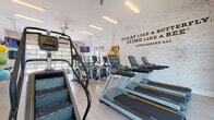 This single-story fitness center is thoughtfully designed to support a full range of workout routines. The space is bright and open, characterized by light wood-look flooring, white-painted brick walls, and high ceilings that contribute to an airy, motivating atmosphere. Natural light pours in generously through multiple windows and a striking floor-to-ceiling glass garage door, which opens toward an outdoor area featuring a pool and surrounding greenery.

The cardio zone is well-appointed with a row of Matrix treadmills and elliptical trainers, each equipped with individual screens. A stair climber adds variety to the cardio lineup. A bold Muhammad Ali quote—"Float like a butterfly, sting like a bee"—is displayed across the white brick accent wall, adding an energetic and inspirational tone to the space. A pair of oversized boxing gloves serves as a decorative focal point on this wall.

The free weight area features a full rack of dumbbells alongside a adjustable weight bench, positioned near the glass garage door with views outdoors. A large wall mirror enhances the sense of space. The functional training area includes a spin bike, stability balls, resistance bands, a yoga mat, and a small beverage refrigerator, making it a versatile space for varied fitness needs. Overhead fluorescent lighting and ceiling fans ensure comfort throughout.