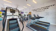 This single-story fitness center is thoughtfully designed to support a full range of workout routines. The space is bright and open, characterized by light wood-look flooring, white-painted brick walls, and high ceilings that contribute to an airy, motivating atmosphere. Natural light pours in generously through multiple windows and a striking floor-to-ceiling glass garage door, which opens toward an outdoor area featuring a pool and surrounding greenery.

The cardio zone is well-appointed with a row of Matrix treadmills and elliptical trainers, each equipped with individual screens. A stair climber adds variety to the cardio lineup. A bold Muhammad Ali quote—"Float like a butterfly, sting like a bee"—is displayed across the white brick accent wall, adding an energetic and inspirational tone to the space. A pair of oversized boxing gloves serves as a decorative focal point on this wall.

The free weight area features a full rack of dumbbells alongside a adjustable weight bench, positioned near the glass garage door with views outdoors. A large wall mirror enhances the sense of space. The functional training area includes a spin bike, stability balls, resistance bands, a yoga mat, and a small beverage refrigerator, making it a versatile space for varied fitness needs. Overhead fluorescent lighting and ceiling fans ensure comfort throughout.