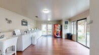 Located at 7100 Shoreline Dr in Stockton, California, this shared laundry facility offers a well-organized, single-floor layout designed for everyday convenience. The main laundry area is lined with a row of top-load washers and dryers along one wall, each equipped with digital payment terminals for a modern, cashless experience. A utility sink is available alongside the machines for added functionality. The space features warm wood-look flooring throughout, cream-colored walls accented with crown molding, and a textured ceiling with flush-mounted lighting that keeps the room bright and functional. A beverage vending machine is conveniently positioned near the entrance, and a laundry cart is on hand for residents' use. Large windows and a set of glass double doors flood the space with natural light and provide views of the landscaped courtyard outside, creating an open and airy atmosphere. A sliding glass door on one side offers an additional connection to the outdoor surroundings. The facility also includes an all-gender restroom with a window for natural light, wood-look flooring, and essential fixtures. Posted signage throughout the facility guides users on best practices and mobile payment options, making this a practical and user-friendly amenity.