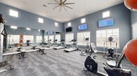 Located at 1640 N Zaragoza Rd in El Paso, TX, this fitness center offers a thoughtfully designed exercise space that caters to a wide range of workout needs. The room is characterized by soaring ceilings that create an open, airy atmosphere, with clerestory windows positioned high on the walls allowing ample natural light to fill the space throughout the day. The walls are finished in a contemporary steel-blue tone, complemented by neutral-toned patterned carpet flooring that adds a modern, polished look.

The cardio zone features multiple treadmills and elliptical machines, as well as an upright stationary bike, all positioned to take advantage of both the natural light from large windows with white blinds and the multiple wall-mounted flat-screen TVs for entertainment during workouts. A full-wall mirror along one side visually expands the room and aids in form correction during exercise.

The strength training area is equally well-appointed, housing a multi-function cable machine, a tiered dumbbell rack with a comprehensive weight selection, adjustable workout benches, and stability balls for functional training. Decorative geometric wall panels add a stylish, gallery-like touch to the strength zone. A large ceiling fan ensures comfortable air circulation, making this fitness center a comprehensive and inviting space for daily training.