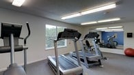 This single-floor fitness room offers a functional workout space equipped with multiple treadmills and cardio machines positioned for ease of use. The room features a textured ceiling with bright fluorescent lighting that ensures clear visibility throughout. A window along one wall allows natural light to filter in, offering a view of surrounding greenery that adds a refreshing element to the workout environment. A large mirror spans a section of the wall, visually expanding the space while allowing users to monitor their form during exercise. The flooring is a durable, low-pile carpet suited for gym use. An accent wall in blue adds a subtle pop of color to the otherwise neutral, light-toned interior. The layout is straightforward and practical, with equipment arranged to maximize available floor space. Overall, the room provides a clean, no-frills environment designed to meet basic cardio fitness needs.