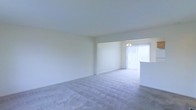 This two-floor residential space offers a straightforward and functional layout designed to maximize usability. The main living area is open and spacious, featuring soft, neutral-toned wall-to-wall carpeting that creates a warm and comfortable underfoot feel. Bright white walls keep the space feeling clean and airy, while a large window dressed with vertical blinds allows natural light to flow generously into the room, enhancing its open atmosphere. A partial dividing wall with a countertop overhang subtly separates the living area from an adjacent dining or kitchen space, maintaining visual connectivity while defining distinct zones. A ceiling-mounted light fixture provides additional illumination to the dining area. The overall design is understated and neutral, offering a versatile canvas well-suited to a variety of interior styles and furnishing choices. The two-floor configuration provides separation between living and private spaces, contributing to a practical and organized flow throughout the home. The consistent use of light tones across flooring, walls, and ceilings gives the entire space a cohesive, bright, and welcoming character.