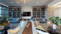 This thoughtfully designed single-floor space blends industrial and contemporary elements to create a versatile, welcoming environment. Exposed brick walls serve as a defining architectural feature throughout, adding warmth and texture that contrasts beautifully with crisp white surfaces and modern finishes.

The kitchen is a standout area, boasting soaring ceilings with exposed beams, a large marble-topped island with adjustable industrial-style bar stools, and a striking trio of antique brass pendant lights. A full-height white tile backsplash, stainless steel appliances, and open shelving complete the space. Floor-to-ceiling glass sliding doors flood the kitchen with natural light and provide direct views of a lush outdoor courtyard.

The lounge areas are equally impressive, featuring built-in gray shelving units flanking a wall-mounted television, comfortable sofas arranged around a rustic wood coffee table, and herringbone hardwood flooring. A floor-to-ceiling glass window wall in one seating area frames outdoor views and draws in abundant natural light.

A dedicated dining area features a large white communal table beneath globe pendant lights, set against an exposed brick wall. A living wall with tiered wooden planters filled with trailing greenery adds a vibrant natural accent throughout the space.
