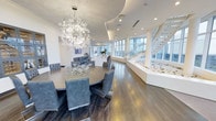 This elegantly designed single-floor space exudes a refined, contemporary aesthetic characterized by clean lines, a neutral white-and-grey palette, and high-end finishes throughout. The open-plan layout allows the interior to flow seamlessly from one area to the next, creating a sense of generous, uninterrupted space.

A standout feature of the dining area is the large, oval dark-wood dining table surrounded by tufted grey upholstered chairs, perfectly complemented by a dramatic, starburst crystal chandelier suspended overhead. The sculptural light fixture serves as a striking focal point, adding a glamorous, artistic touch to the room.

Warm hardwood flooring extends throughout the space, grounding the interior with an organic texture that contrasts beautifully against the bright white walls and ceiling. The curved, tiered ceiling detailing adds architectural depth and visual interest.

Along one side of the space, expansive floor-to-ceiling windows flood the interior with abundant natural light and offer panoramic views of the surrounding cityscape. Adjacent to the windows, a sleek floating staircase with glass balustrades ascends gracefully, serving as both a functional element and a sculptural architectural statement. A decorative rock garden at the base of the staircase introduces a natural, serene element to the otherwise polished interior.
