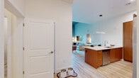 This single-floor residence showcases a thoughtfully designed open-concept layout where the kitchen, dining, and living areas flow seamlessly together. The kitchen is a standout feature, equipped with flat-front wood cabinetry, stainless steel appliances including a French door refrigerator, a tile backsplash, and a generous island with a white quartz countertop and seating for two. A linear pendant light anchors the island, complementing the recessed lighting overhead.

Light wood-tone flooring runs throughout the entire residence, providing a warm, cohesive foundation. The living area is distinguished by dramatically tall ceilings and a striking exposed concrete accent wall that adds an industrial character. Sliding glass doors open directly to a private balcony with outdoor views, filling the space with natural light.

The bedroom continues the concrete accent wall detail, reinforcing the unit's cohesive design language, while soaring ceilings amplify the sense of spaciousness. A dedicated closet area with built-in shelving provides organized storage adjacent to the bedroom.

The bathrooms feature wood-toned vanities, tile-surround shower and tub combinations, and light wood flooring, maintaining the modern and clean aesthetic found throughout the residence.
