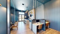 Located at 208 Windfall Way in Myrtle Beach, SC, this thoughtfully designed single-floor amenity space delivers a coastal-inspired atmosphere through a cohesive blend of bold color, natural materials, and functional layout. The interior is anchored by deep navy blue walls and a matching painted ceiling that create a rich, immersive backdrop throughout the room. Light oak hardwood flooring runs continuously across the space, providing warmth and contrast against the cooler tones above.

A central white-topped bar counter with dark metal stools serves as the social hub of the room, flanked by vibrant teal cabinetry and a white geometric tile backsplash. A wall-mounted flat-screen TV and open wood shelving with curated décor add visual interest to this refreshment and gathering area. A striking installation of suspended white ropes cascades from the ceiling alongside teardrop-shaped metallic pendant lights, adding a distinctly nautical design accent.

Dedicated workstations with built-in desks and caramel leather chairs are neatly tucked into a partitioned zone, offering a quiet and focused environment. At the rear of the space, a windowed alcove with bold yellow-and-white patterned wallpaper, built-in bench seating, and small round tables creates an inviting nook filled with natural light and outdoor views. French doors at the entry allow additional light to pour in, reinforcing the open and airy character of this well-appointed community space.
