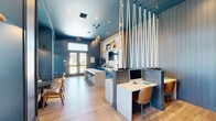 Located at 208 Windfall Way in Myrtle Beach, SC, this thoughtfully designed single-floor amenity space delivers a coastal-inspired atmosphere through a cohesive blend of bold color, natural materials, and functional layout. The interior is anchored by deep navy blue walls and a matching painted ceiling that create a rich, immersive backdrop throughout the room. Light oak hardwood flooring runs continuously across the space, providing warmth and contrast against the cooler tones above.

A central white-topped bar counter with dark metal stools serves as the social hub of the room, flanked by vibrant teal cabinetry and a white geometric tile backsplash. A wall-mounted flat-screen TV and open wood shelving with curated décor add visual interest to this refreshment and gathering area. A striking installation of suspended white ropes cascades from the ceiling alongside teardrop-shaped metallic pendant lights, adding a distinctly nautical design accent.

Dedicated workstations with built-in desks and caramel leather chairs are neatly tucked into a partitioned zone, offering a quiet and focused environment. At the rear of the space, a windowed alcove with bold yellow-and-white patterned wallpaper, built-in bench seating, and small round tables creates an inviting nook filled with natural light and outdoor views. French doors at the entry allow additional light to pour in, reinforcing the open and airy character of this well-appointed community space.