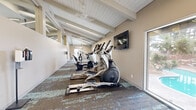 Located in Huntington Beach, California, this well-appointed fitness center offers a spacious, single-level layout designed to support a full range of exercise activities. The room is defined by a striking vaulted ceiling with exposed white-painted beams and integrated linear LED lighting that runs the length of the space, creating a bright and energizing atmosphere throughout.

The fitness floor is covered in a multi-toned, textured carpet that adds visual interest while providing comfortable underfoot support. Large sliding glass doors and generous windows line one side of the room, flooding the interior with natural light and offering direct views of an outdoor pool and landscaped surroundings — a motivating backdrop for any workout.

Equipment is thoughtfully arranged along the length of the room and includes elliptical trainers, treadmills, a recumbent bike, and a multi-function strength training station. Wall-mounted flat-screen televisions provide entertainment throughout the space. A dedicated open area near the pool-facing windows accommodates flexibility and stretching, and is stocked with stability balls, exercise mats, and a water station.

The entry area features a glass-paneled door with transom windows, accent stone tile detailing, and a small seating nook with lounge chairs — offering a welcoming transition into the fitness space.