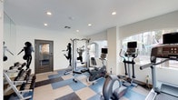 This single-level fitness room offers a clean, functional space designed to support a well-rounded workout routine. The room features a multi-station cable machine as its centerpiece, complemented by cardio equipment including an elliptical trainer and an upright stationary bike, each equipped with integrated display monitors. The flooring consists of a blue and gray checkered tile pattern that adds a dynamic visual element while providing a durable, easy-to-clean surface suited for physical activity. Recessed lighting in the white ceiling ensures consistent, even illumination throughout the space. The walls are finished in a neutral white tone, contributing to the room's bright and open atmosphere. Decorative silhouette decals of athletes in motion are applied to the walls, reinforcing the fitness-oriented character of the room. Natural light is introduced through windows and a glass-paned door, offering views of lush outdoor greenery and creating an airy ambiance. An arched architectural detail adds a subtle design element that breaks the room's otherwise straightforward layout. Overall, the space is efficiently organized, providing accessible equipment and a motivating environment for exercise.