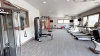 Located at 21900 East Country Vista Drive in Liberty Lake, Washington, this single-level fitness center offers a comprehensive and thoughtfully arranged workout facility. The space is organized into distinct training zones, each designed to support a wide range of fitness activities.

The cardio area is lined with True-brand treadmills, elliptical trainers, and stationary bikes, positioned along large windows and an impressive glass-panel garage door that opens fully to the outdoors. This feature floods the space with natural light and fresh air while offering views of the surrounding grounds.

The strength training section houses multiple Inspire Fitness dual cable machines with adjustable benches, providing versatile resistance training options. A dedicated functional training zone features a bold red power rack with pull-up bars, TRX suspension trainers, battle ropes, a full dumbbell rack, kettlebells, medicine balls, and a stability ball — accommodating everything from high-intensity interval training to mobility work.

Throughout the facility, durable patterned carpet tiles cover the floors, and recessed lighting ensures consistent illumination. Wall-mounted televisions are positioned throughout the space for entertainment during workouts. The clean white walls, warm wood-toned trim accents, and organized equipment layout contribute to a modern, functional, and welcoming fitness environment.