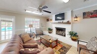 Located at 2998 Ross Clark Circle in Dothan, Alabama, this single-floor space offers a thoughtfully designed combined living and dining area that maximizes its compact footprint. The living room is anchored by a striking white-painted brick fireplace with a rustic wood mantel, above which a wall-mounted television is positioned for easy viewing. A ceiling fan with a light fixture overhead provides both comfort and illumination, while crown molding and a textured ceiling add architectural detail throughout. The warm, neutral-toned walls are complemented by tile flooring, creating a bright and inviting atmosphere. A large sliding glass door in the dining area floods the space with natural light and offers direct views of the outdoor pool area, seamlessly connecting the interior to the exterior. The dining space is accessed through a charming French door entrance and features a compact refrigeration unit, adding practical utility. Outside, the property is enclosed by classic brick and wrought iron fencing, surrounding a well-maintained pool deck lined with lounge chairs beneath mature shade trees — an ideal setting for relaxation. The brick exterior architecture and lush landscaping contribute to the space's warm, residential character.