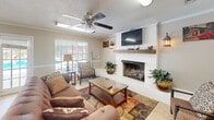 Located at 2998 Ross Clark Circle in Dothan, Alabama, this single-floor space offers a thoughtfully designed combined living and dining area that maximizes its compact footprint. The living room is anchored by a striking white-painted brick fireplace with a rustic wood mantel, above which a wall-mounted television is positioned for easy viewing. A ceiling fan with a light fixture overhead provides both comfort and illumination, while crown molding and a textured ceiling add architectural detail throughout. The warm, neutral-toned walls are complemented by tile flooring, creating a bright and inviting atmosphere. A large sliding glass door in the dining area floods the space with natural light and offers direct views of the outdoor pool area, seamlessly connecting the interior to the exterior. The dining space is accessed through a charming French door entrance and features a compact refrigeration unit, adding practical utility. Outside, the property is enclosed by classic brick and wrought iron fencing, surrounding a well-maintained pool deck lined with lounge chairs beneath mature shade trees — an ideal setting for relaxation. The brick exterior architecture and lush landscaping contribute to the space's warm, residential character.