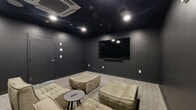 Located at 269 Orange Street in New Haven, CT, this dedicated screening room offers a compact yet immersive cinematic experience. The space is defined by its dramatic all-dark aesthetic, with deep charcoal walls, a near-black ceiling, and neutral-toned carpet that together create a focused, theater-like atmosphere designed to minimize distraction and enhance viewing.

A large wall-mounted flat-screen television serves as the room's centerpiece, paired with a soundbar positioned directly beneath it for an enhanced audio experience. Recessed ceiling lighting and an integrated ceiling-mounted HVAC unit provide both ambiance and climate control, ensuring year-round comfort. In-ceiling speakers are also present, further contributing to an enveloping audio environment.

The seating arrangement features a collection of tufted, modular lounge chairs and ottomans in a warm beige-gray velvet upholstery, offering flexible configurations to suit different group sizes. Small round side tables are distributed throughout the space, providing convenient surfaces for remotes and personal items. The overall layout is thoughtfully intimate, allowing every seat a clear sightline to the screen. This screening room presents a well-appointed, purposefully designed retreat for private viewing and entertainment.